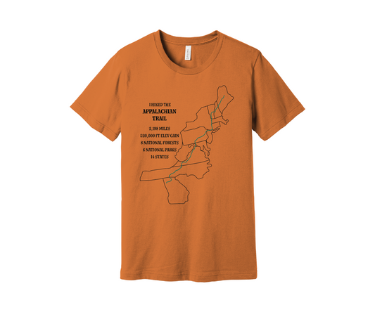 A muted orange T-shirt displays a map outline in black of the Appalachian Trail with a green line marking the route. Text on the shirt reads: "I Hiked the Appalachian Trail," and lists details: 2,198 miles, 464,500 ft elevation gain, 8 National Forests, 6 National Parks, 14 states.