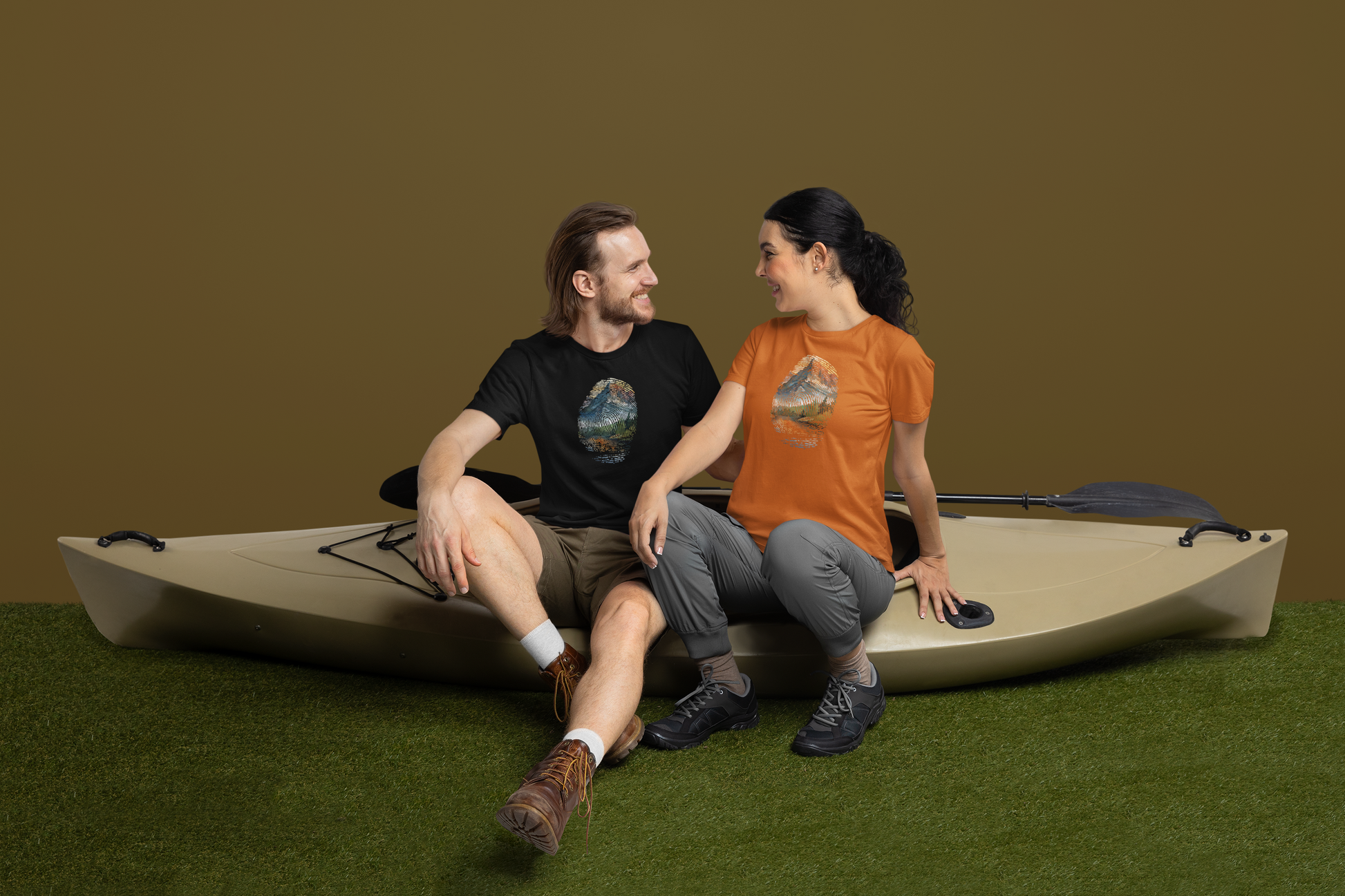 A man and woman sit on a beige kayak inside a room with a brown wall and green floor. The man, in a black t-shirt and khaki shorts, smiles at the woman, who is in an orange t-shirt and gray pants. Both shirts have a mountain graphic encapsulated within a fingerprint. A paddle rests on the kayak.